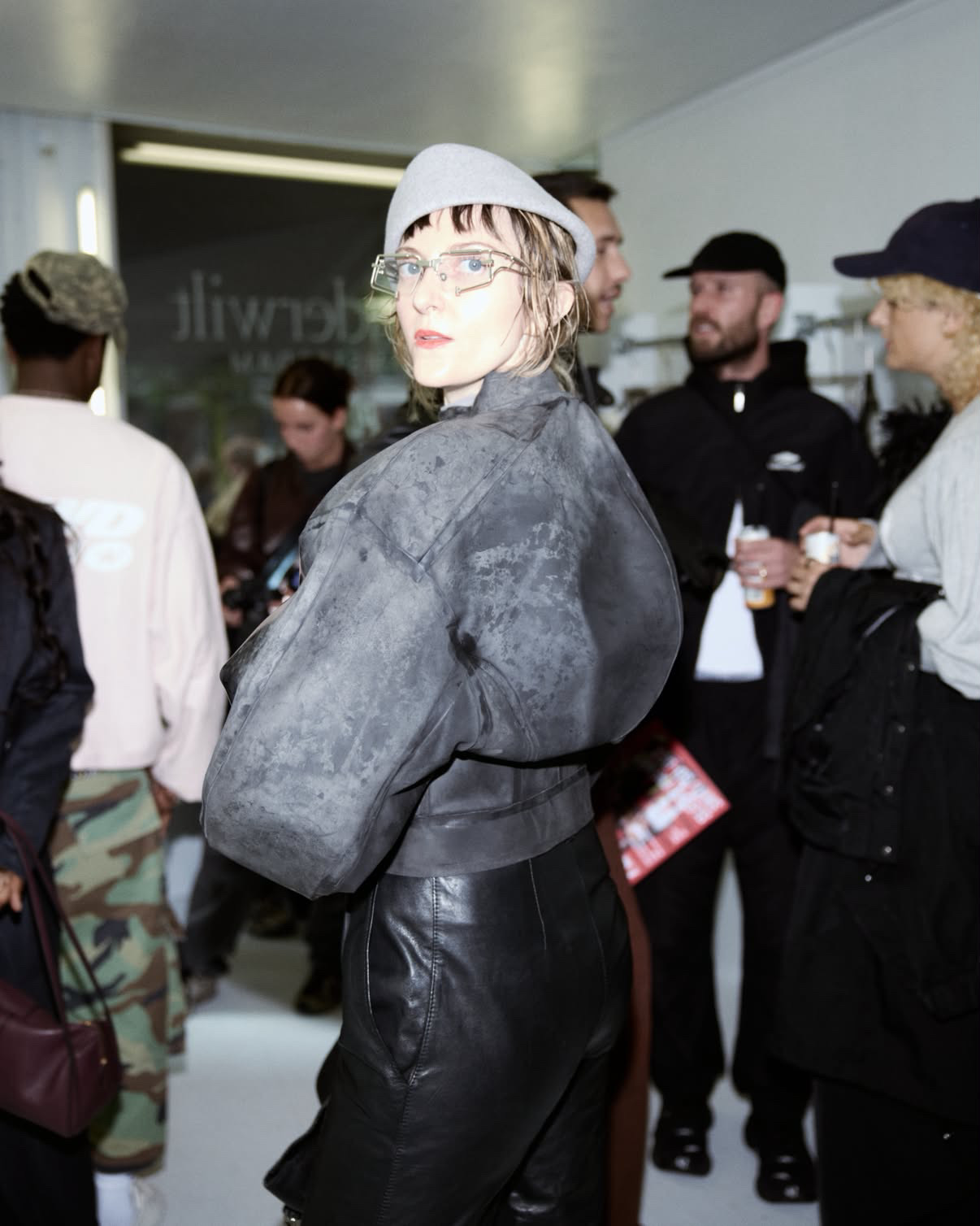 Susan Isko in a grey leather jacket and beret, glancing back at the camera at a fashion event.
